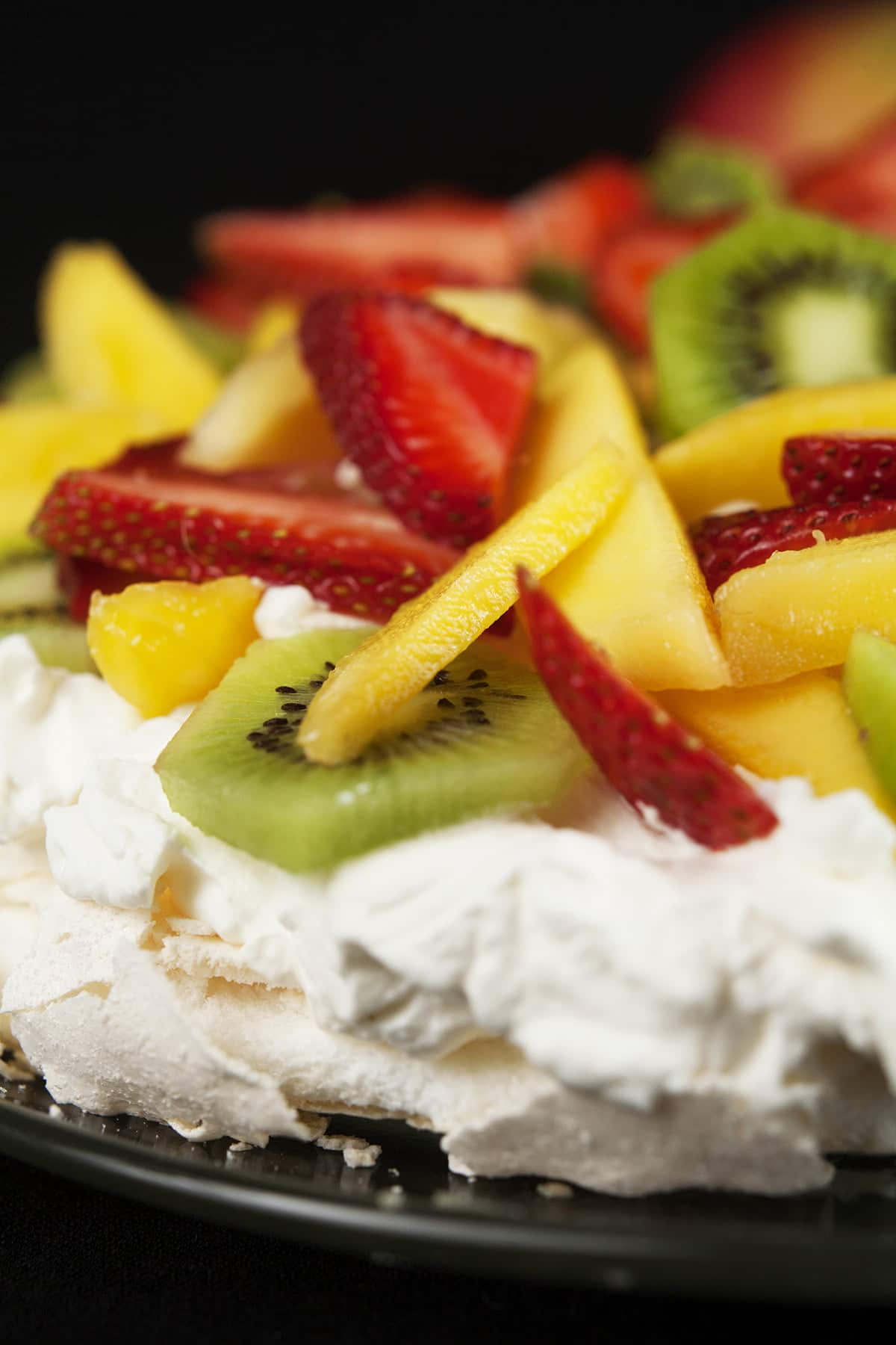 Close up view of a Pavlova: White meringue topped with whipped cream, strawberries, kiwi fruit, and mango slices.