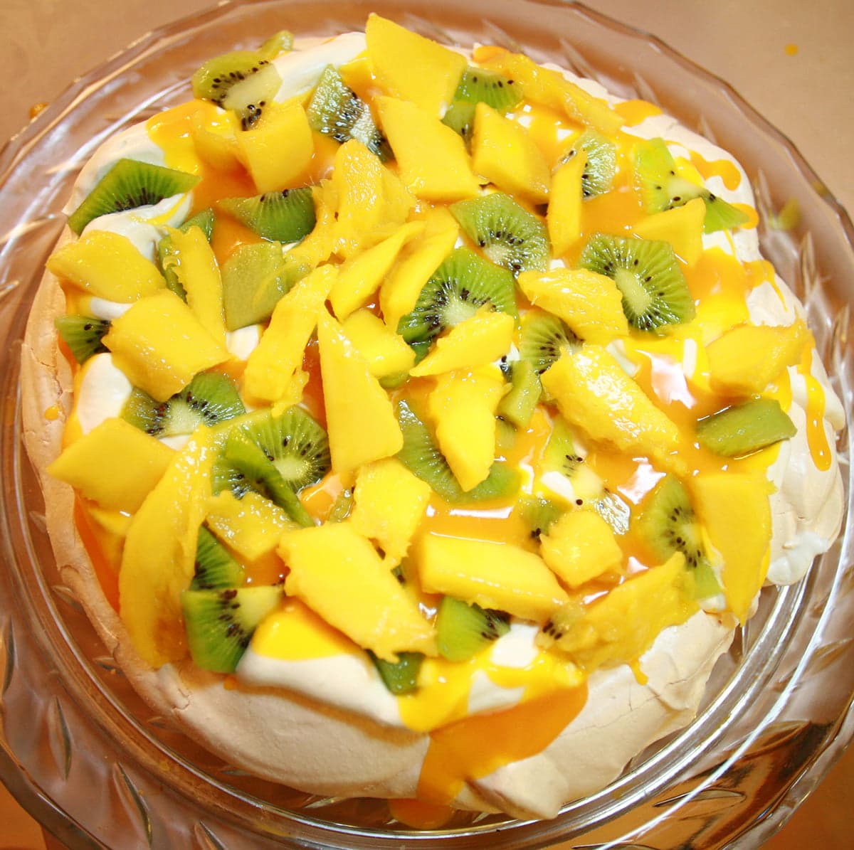 A round white meringue topped with whipped cream, mango slices, kiwi slices, and mango sauce.