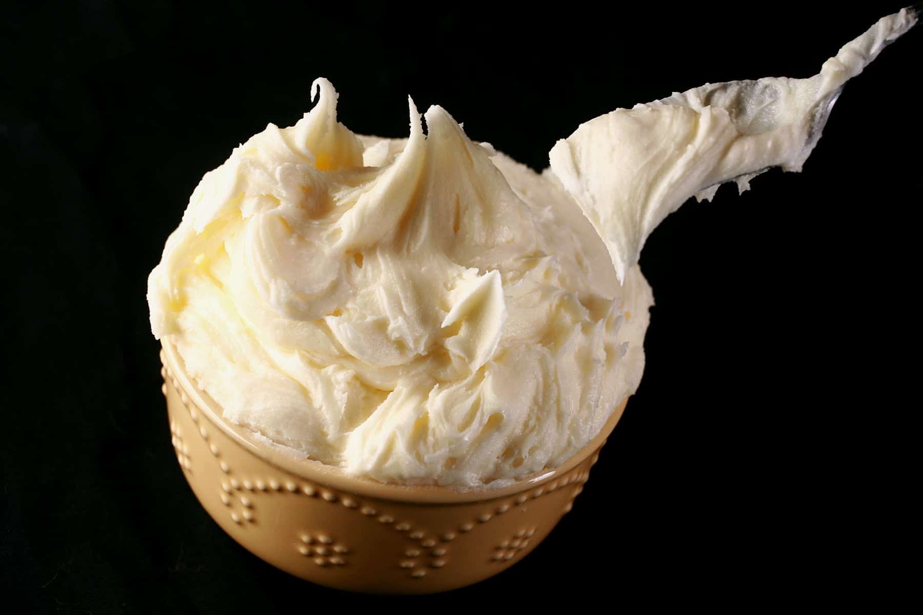 A bowl full of - and mounded high with - off white American Buttercream is shown in front of a black background.