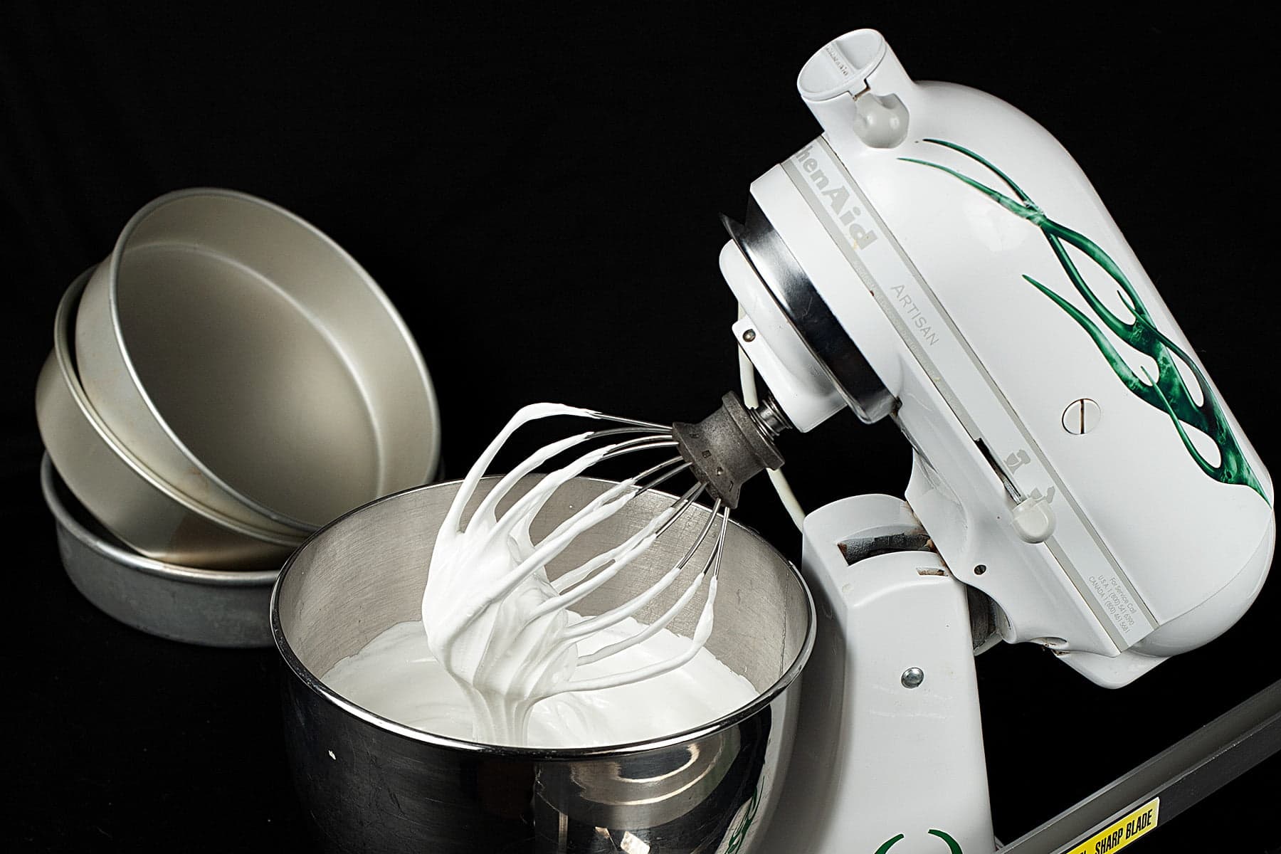 A white stand mixer is pictured against a black background. It's tilted up, showing the meringue in the bowl and on the whisk attachment.