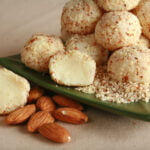 A close up view of a pile of white chocolate truffles, on a small green plate. The truffles are covered in finely chopped almonds, and there are a few whole almonds in the foreground.