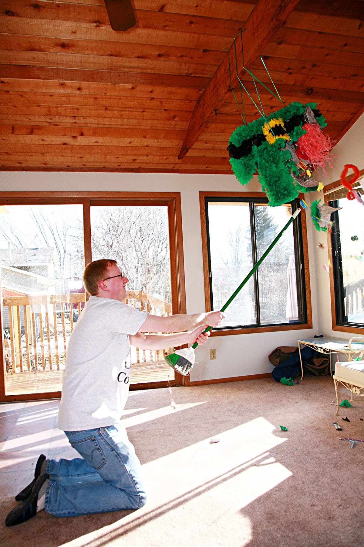 A young blond man wearing a t shirt with a white and black cow print pi symbol on it hits a pinata with a stick.