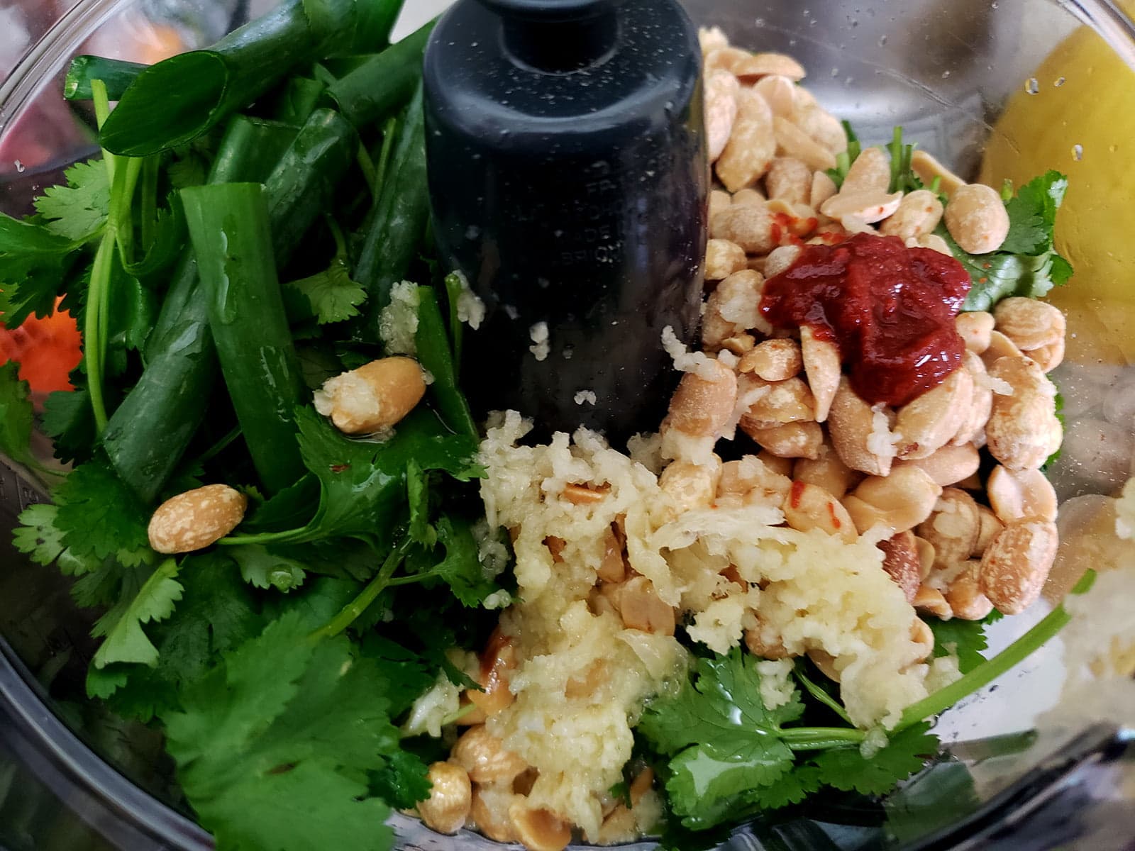 Cilantro, peanuts, garlic, green onions, and sriracha are visible in a mini food processor, waiting to be chopped together.
