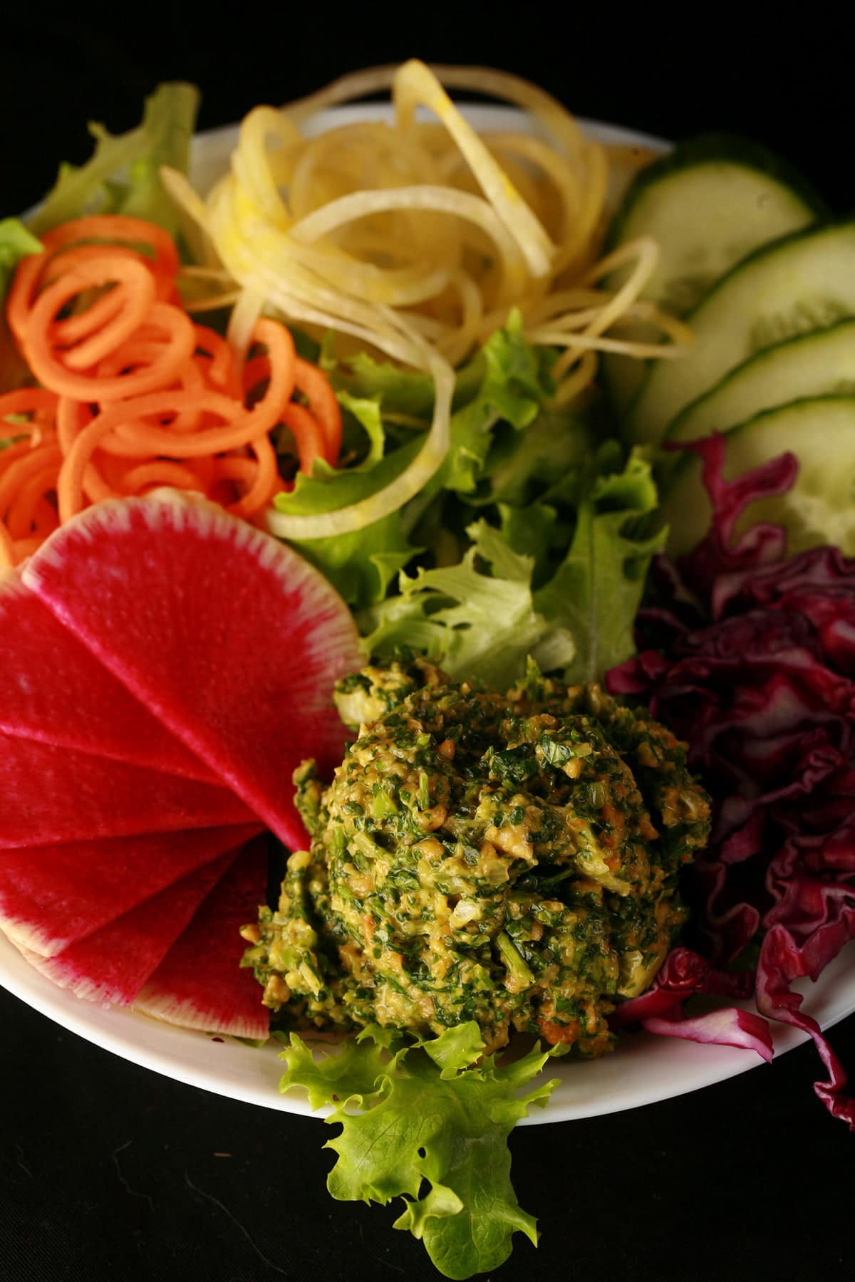 A large scoop of cilantro pesto is shown against the backdrop of a gloriously colourful salad - greens, watermelon radish, spiralized carrots and yellow beets, cucumber, and purple cabbage!