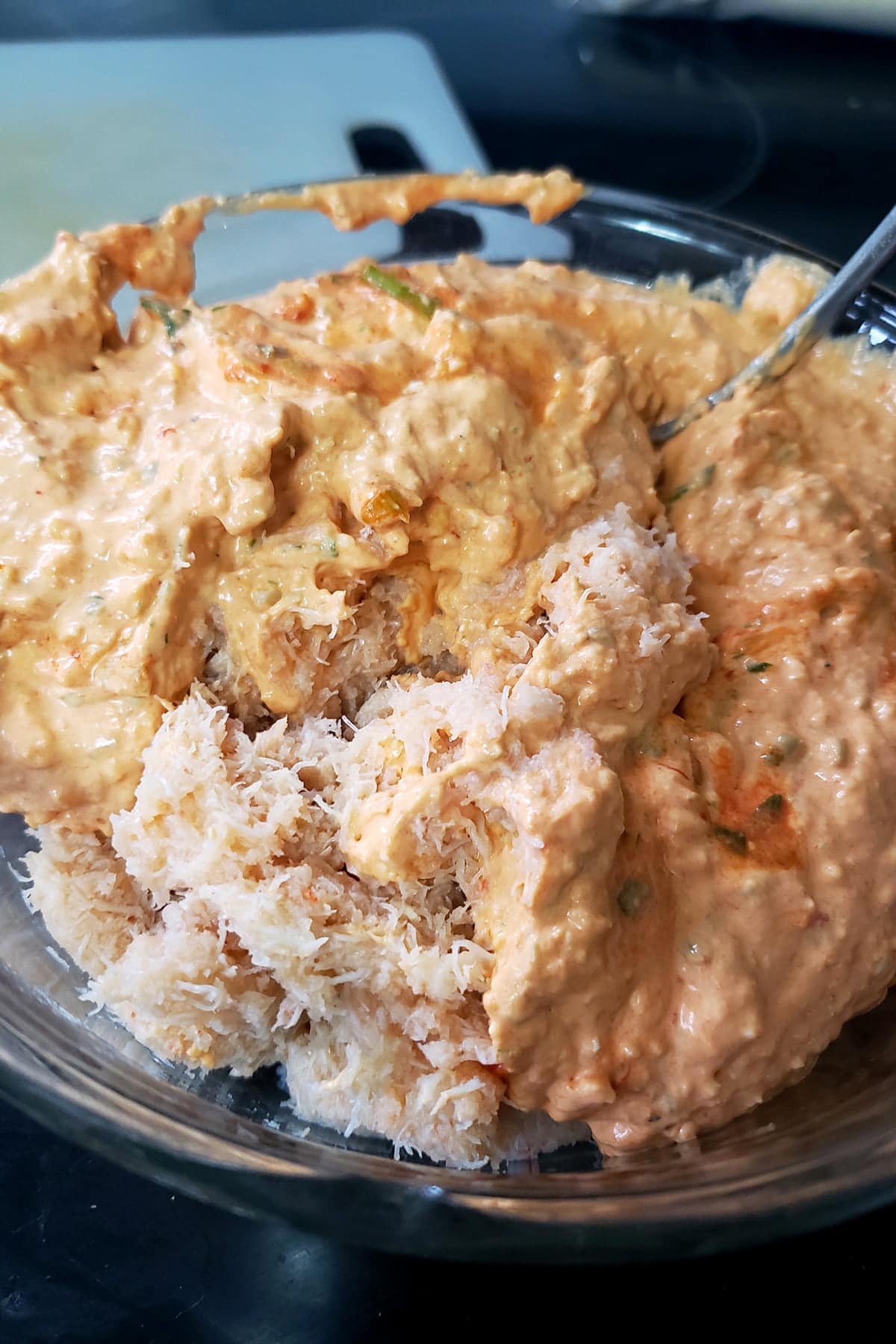 A glass mixing bowl is shown. The red cream cheese mixture has been added to drained crab, and is about to be mixed.