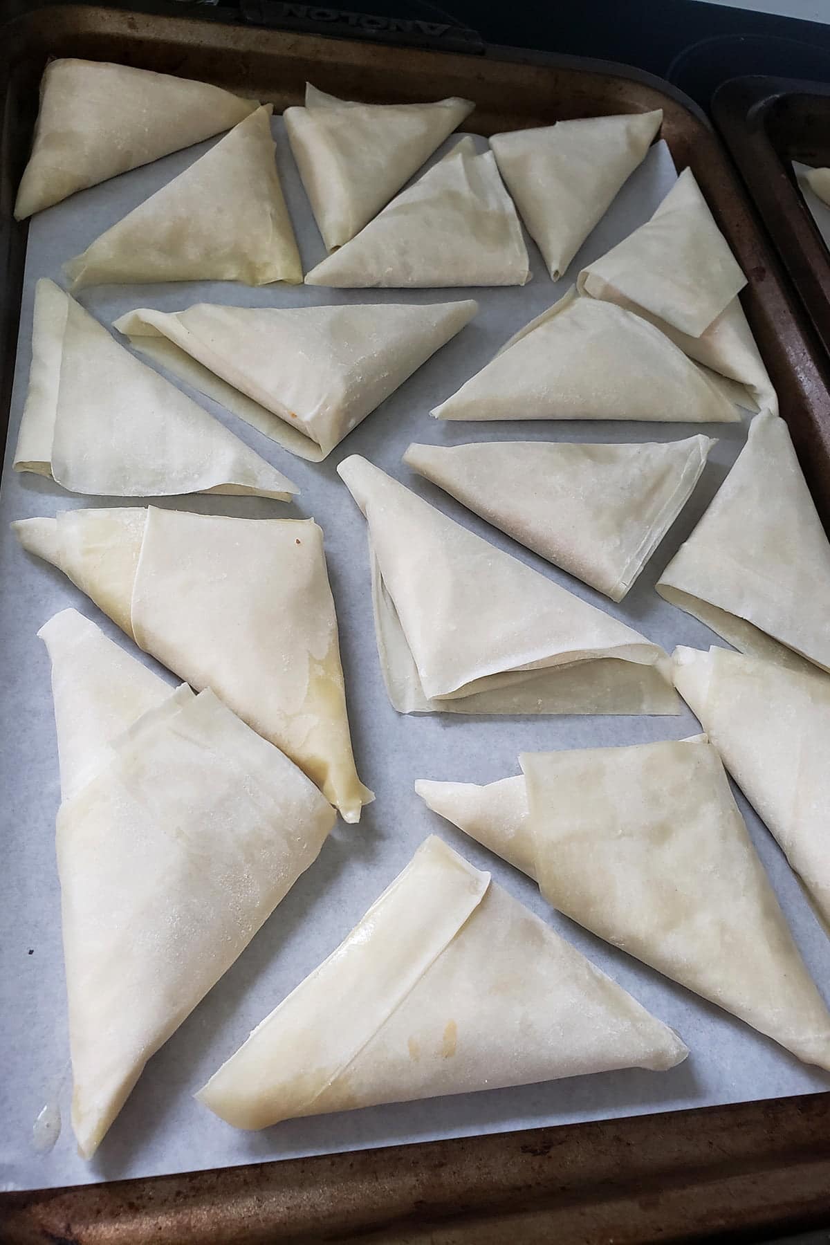 A baking sheet that is lined with parchment paper. About a dozen raw, trianglar pastries have been arranged on the pan, about to be baked.