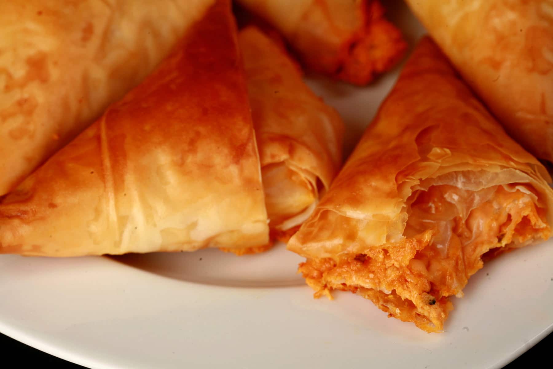 A close up view of crab triangles - a triangular pastry made from folded phyllo dough. They are flaky and crispy, with a red filling showing from one that has been bitten into.