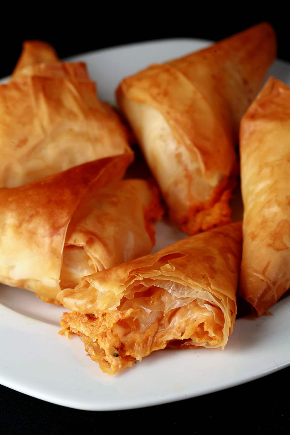 A close up view of a triangular pastry made from folded phyllo dough. They are flaky and crispy, with a red filling showing from one that has been bitten into.