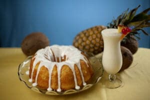A pina colada bundt cake with white glaze dripping from the top is shown next to a tall tulip shaped glass of pina colada. A coconut and pineapple are in the background.