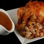 Close up image of a roasted turkey, with an abundance of stuffing visible. It is on a white plate, and there is a bowl of gravy next to it.