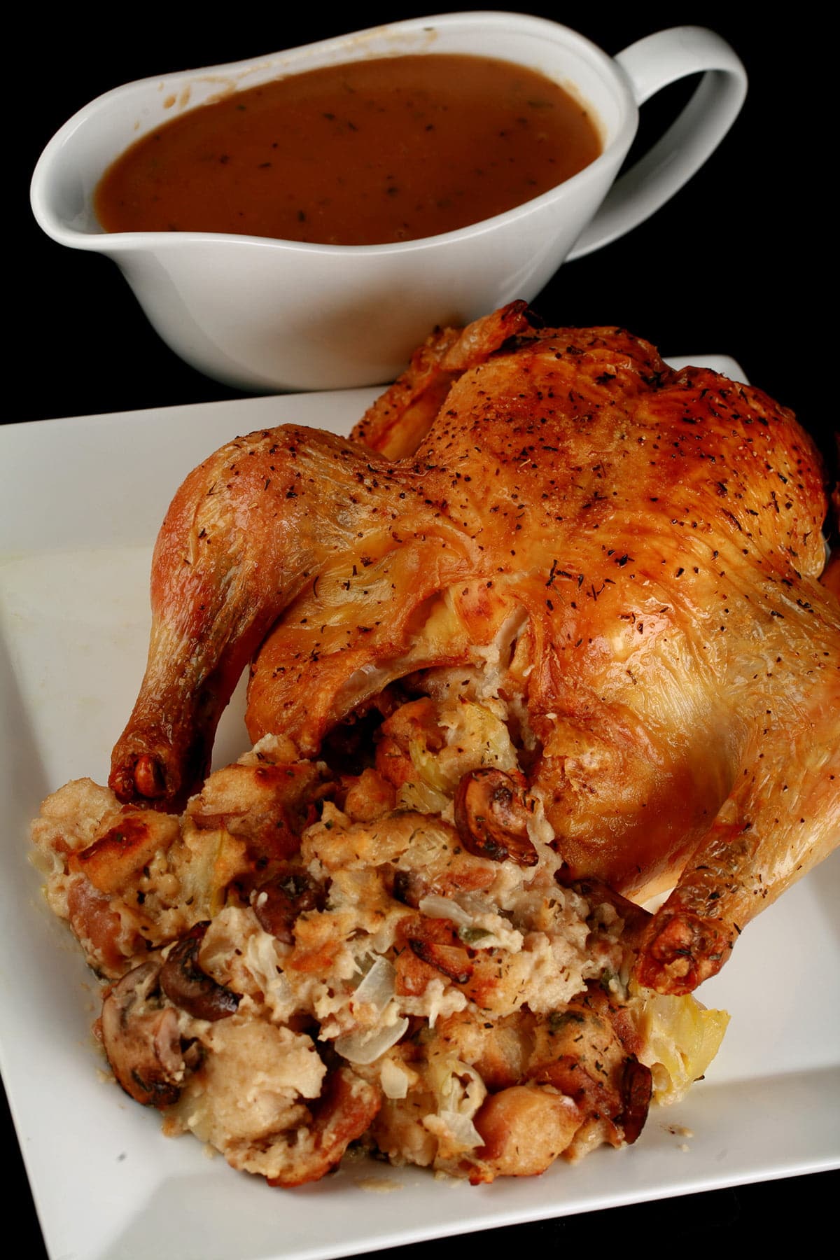 Close up image of a roasted turkey, with an abundance of stuffing visible. It is on a white plate, and there is a bowl of gravy next to it.