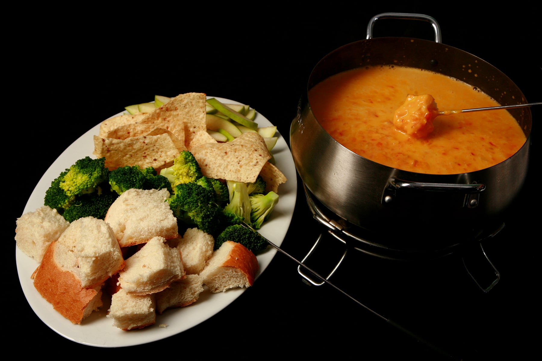 A large platter of bread cubes, broccoli, torilla chips, and apple slices. It's next to a metal fondue pot full of southwest fondue.