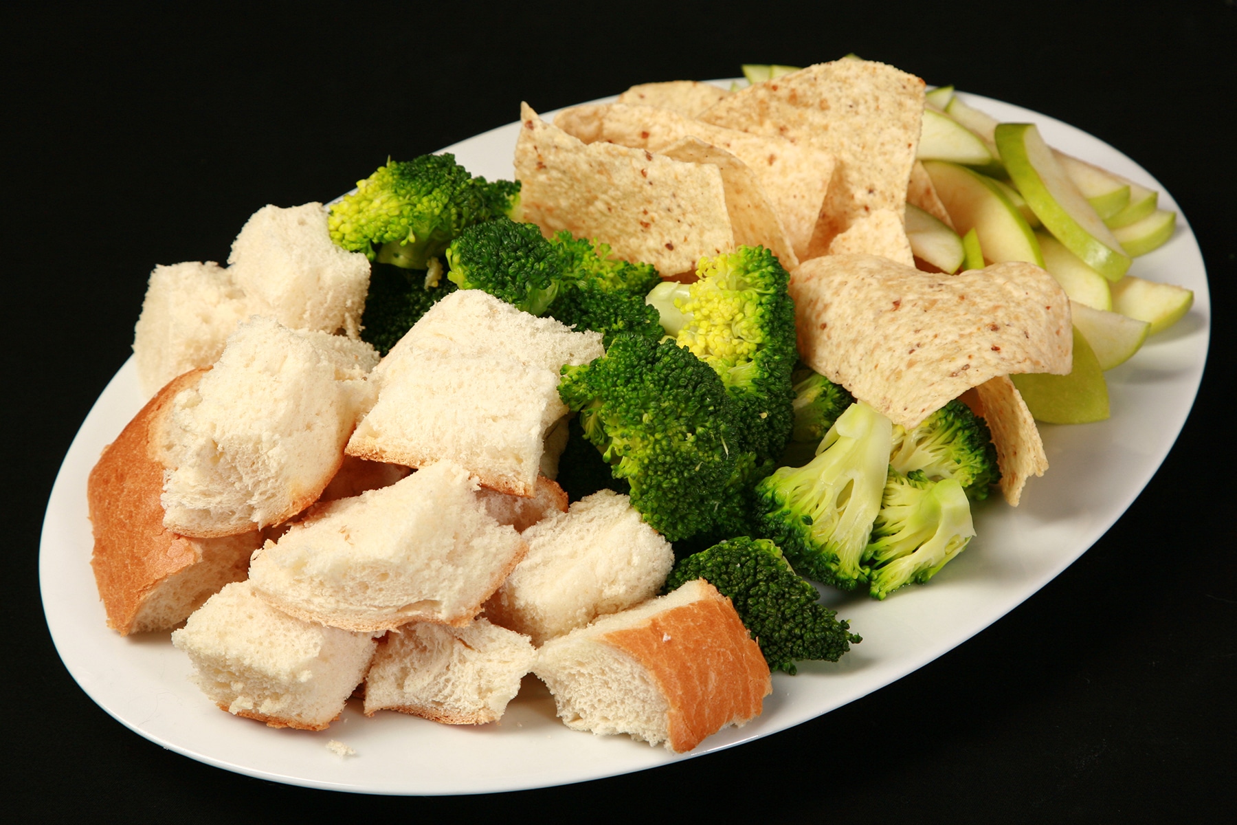 A large platter of bread cubes, broccoli, torilla chips, and apple slices.