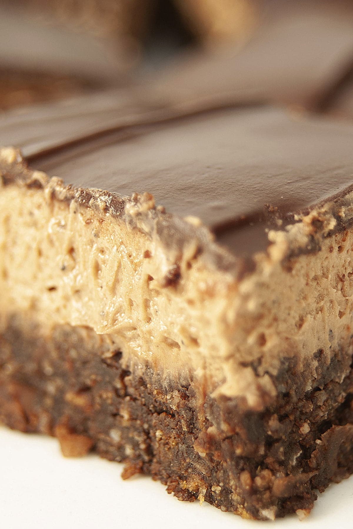 Close up view of a plate of 3 layered bars. The bottom layer looks like a brownie, the middle is a thick brown buttercream, and the top is a smooth chocolate ganache.