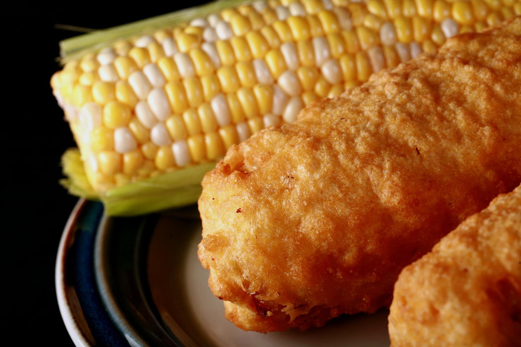 3 cobs of corn - 1 fresh, 2 battered and deep fried - are lined up on a plate.