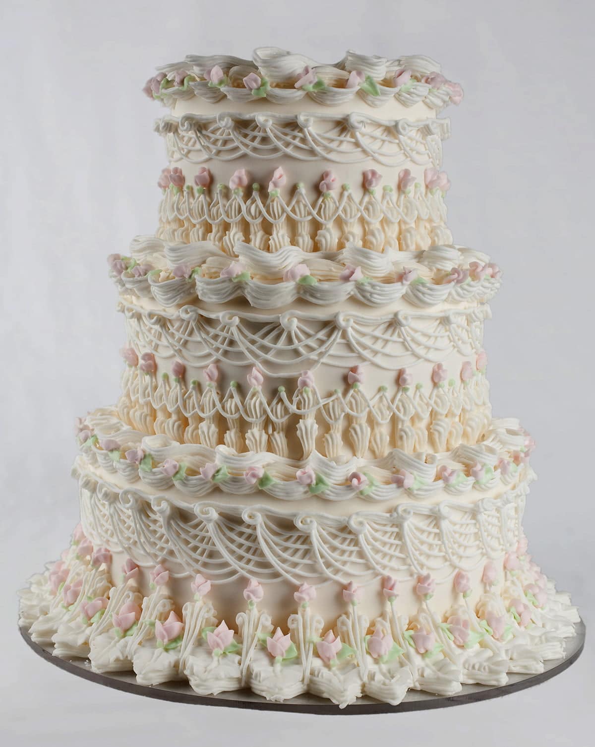 An ornately piped wedding cake, done in the lambeth style. Each layer is adorned with many layers of piping, overpiping, swags, scallops, and more. The cake is mostly white, with pale pink and green accents in the form of rosebuds.