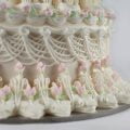 An ornately piped wedding cake, done in the lambeth style. Each layer is adorned with many layers of piping, overpiping, swags, scallops, and more. The cake is mostly white, with pale pink and green accents in the form of rosebuds.
