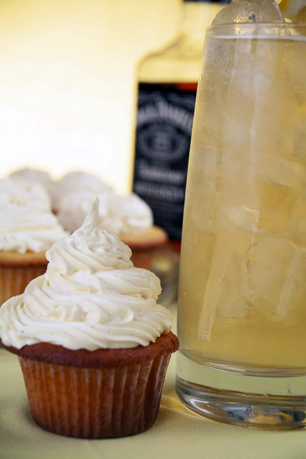 A tray of Lynchburg Lemonade Cupcakes is pictured with a bottle of Jack Daniels, a couple fresh lemons, and a glass of Lynchburg lemonade cocktail.