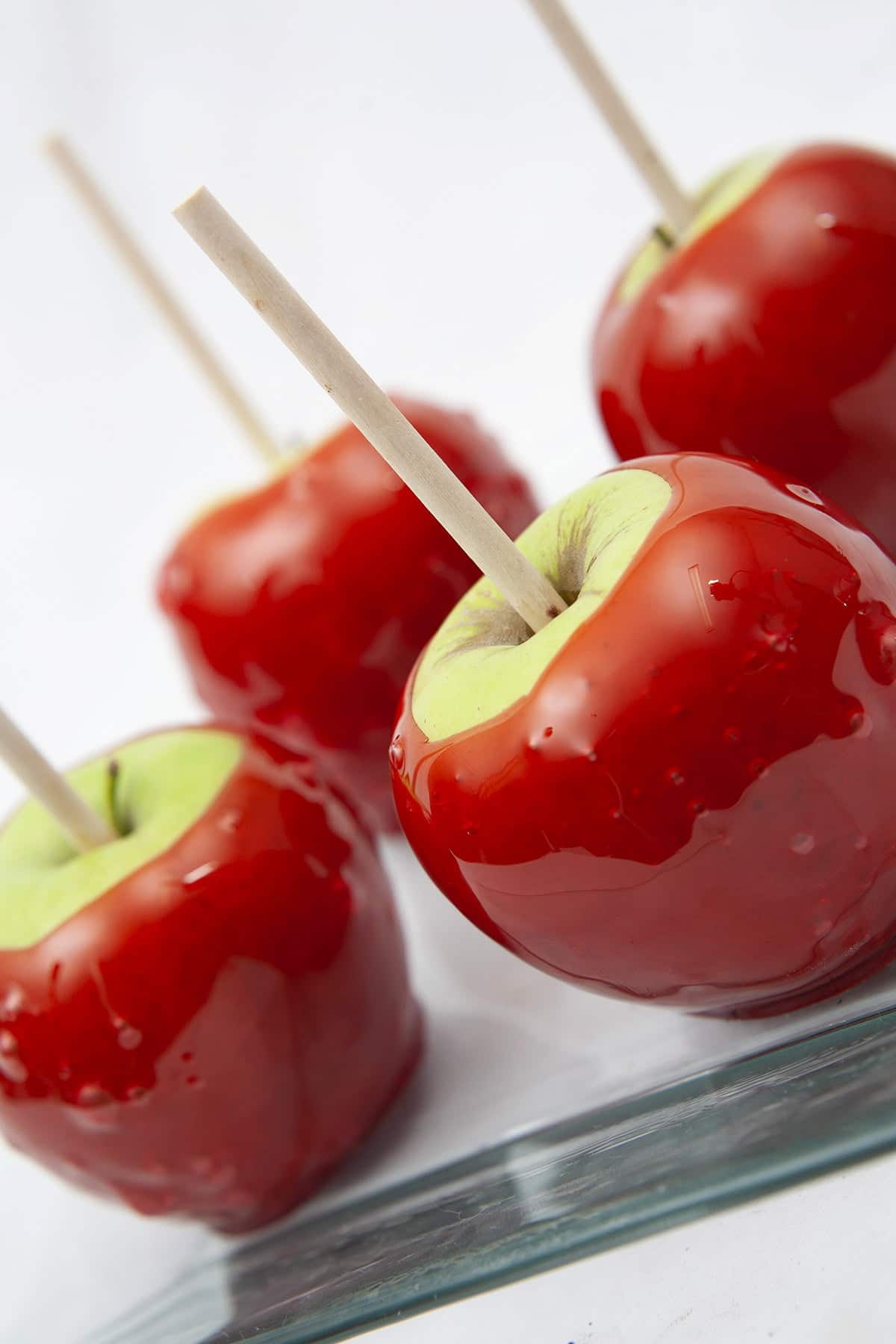 A square shaped glass plate with 4 Candy Apples on it. The apples are green - Granny Smith - and the candy dip on them is a bright shiny red.