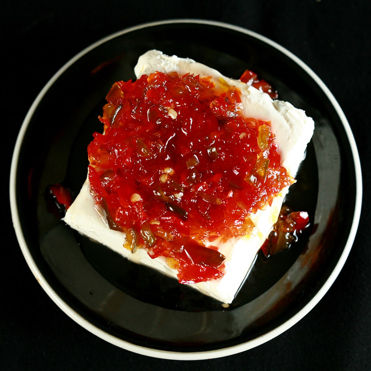 A mound of pepper jelly on a chunk of cream cheese on a small black plate.