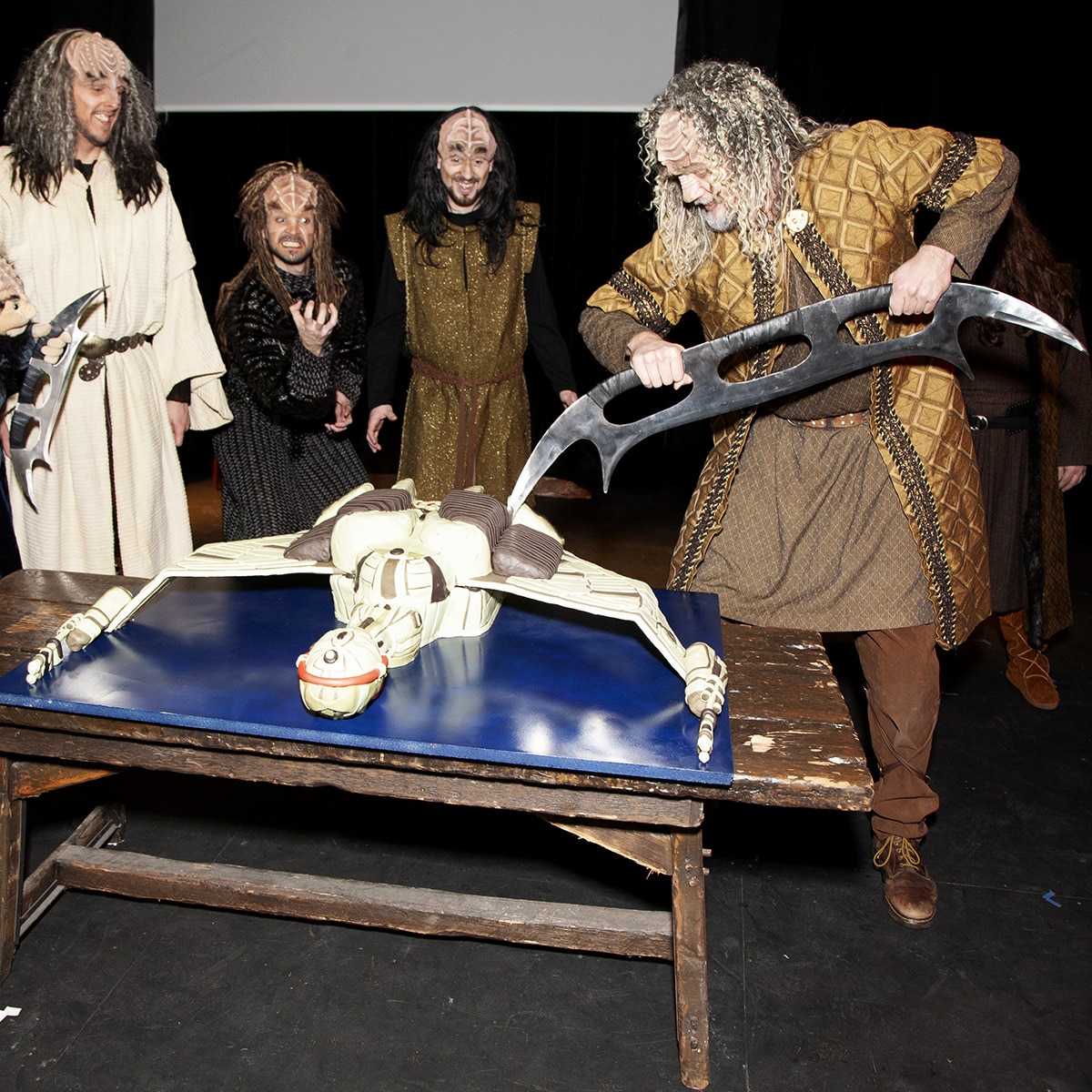 A group of Klingons gathered around a table. on the table is the Klingon ship cake. One Klingon uses his Bat'leth to cut the cake.