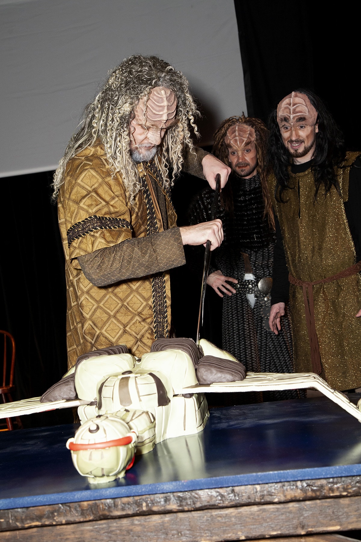 A group of Klingons gathered around a table. on the table is the Klingon ship cake. One Klingon uses his Bat'leth to cut the cake.