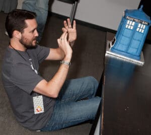 Wil Wheaton under a table, taking a photo of the TARDIS Cake.