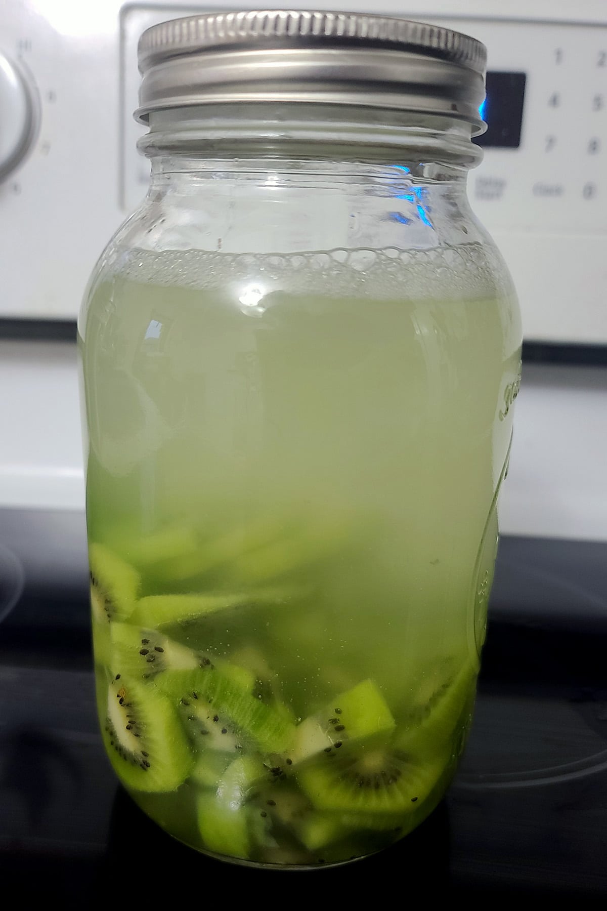 Chopped kiwifruit in a large mason jar with vodka in it. The vodka is getting cloudy.