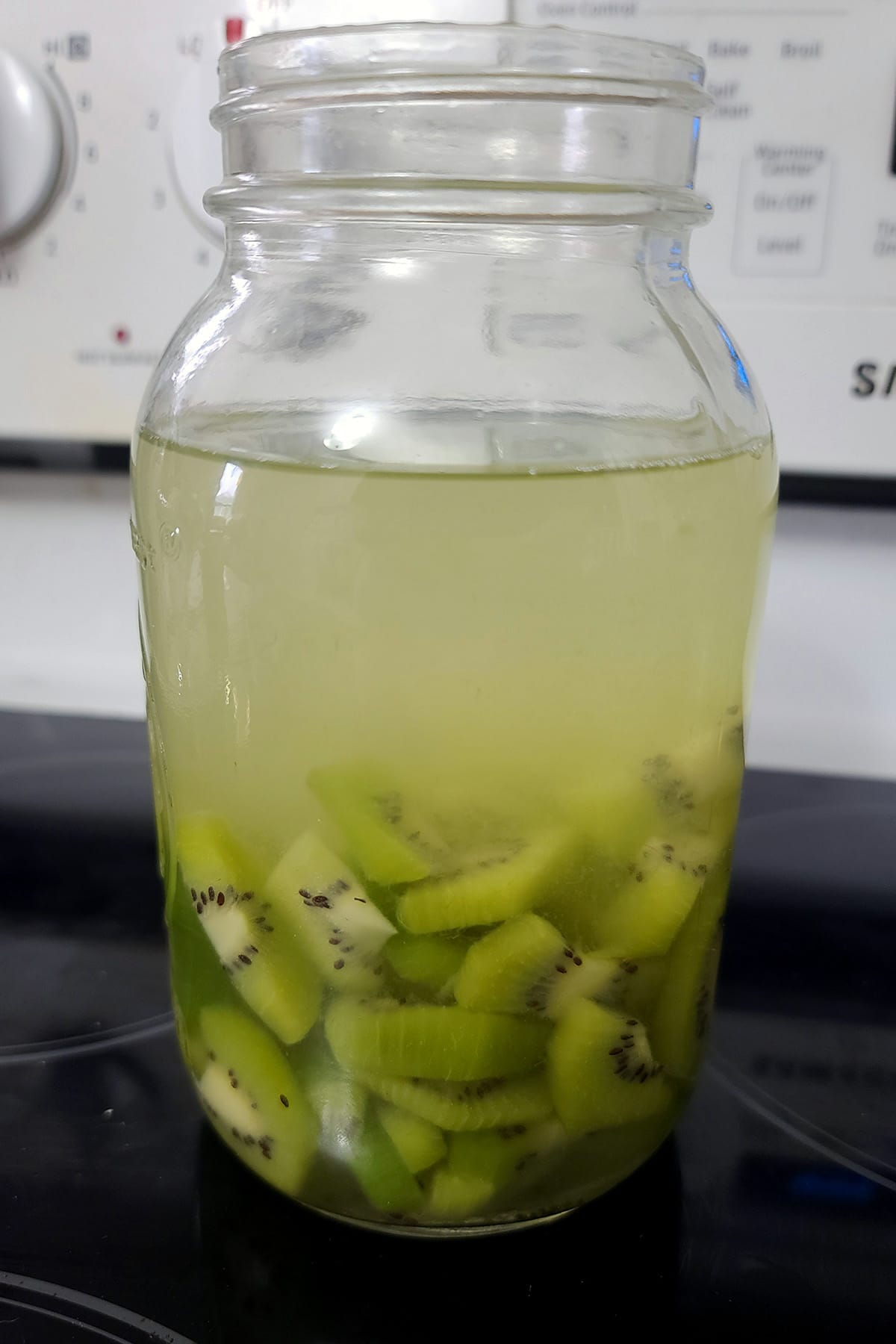 Chopped kiwifruit in a large mason jar with vodka in it. The vodka has a yellow green tinge.