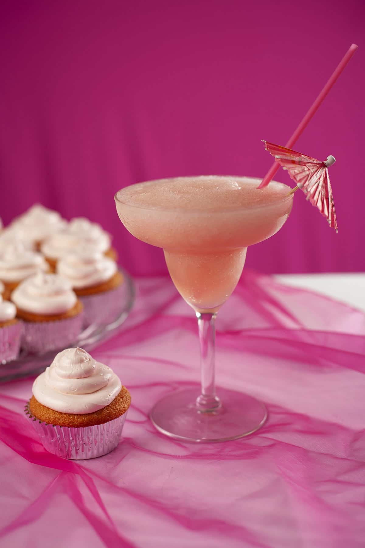 A tray of pink grapefruit daiquiri cupcakes - in silver liners, topped with pink frosting. There is a pink grapefruit daiquiri next to the cupcakes.