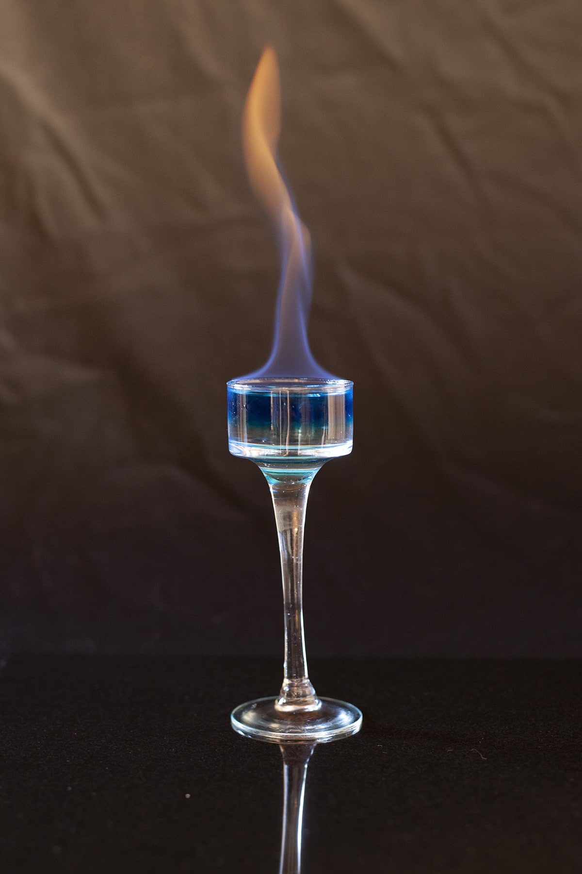 A clear and blue layered shot in a tall, stemmed shot glass. The drink it on fire.