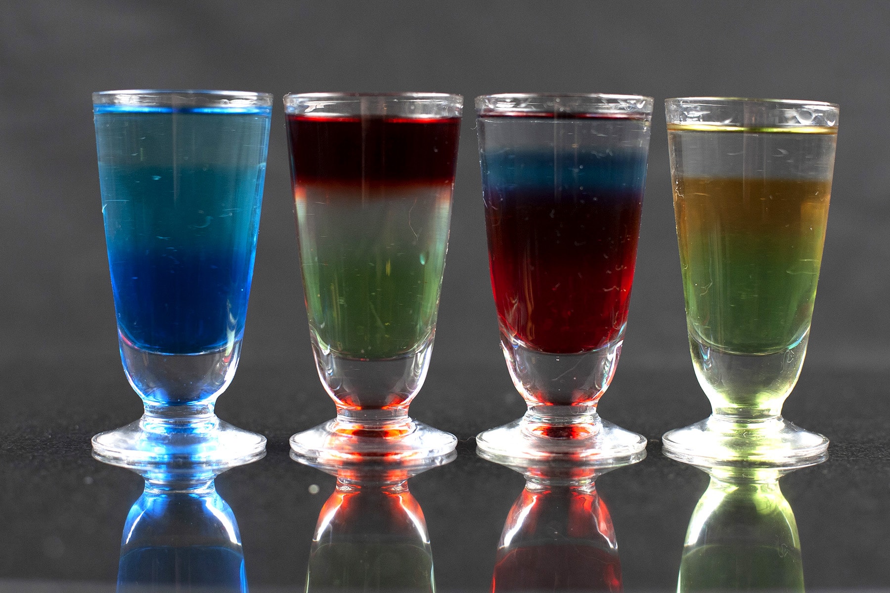 A line of multicoloured layered shooters in stemmed shot glasses.