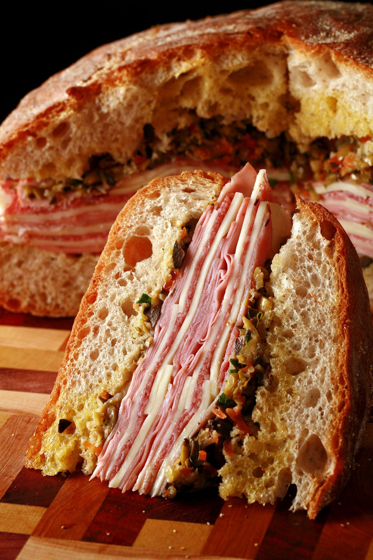 A close up view of a wedge of New Orleans Muffuletta, with the whole sandwich behind it.