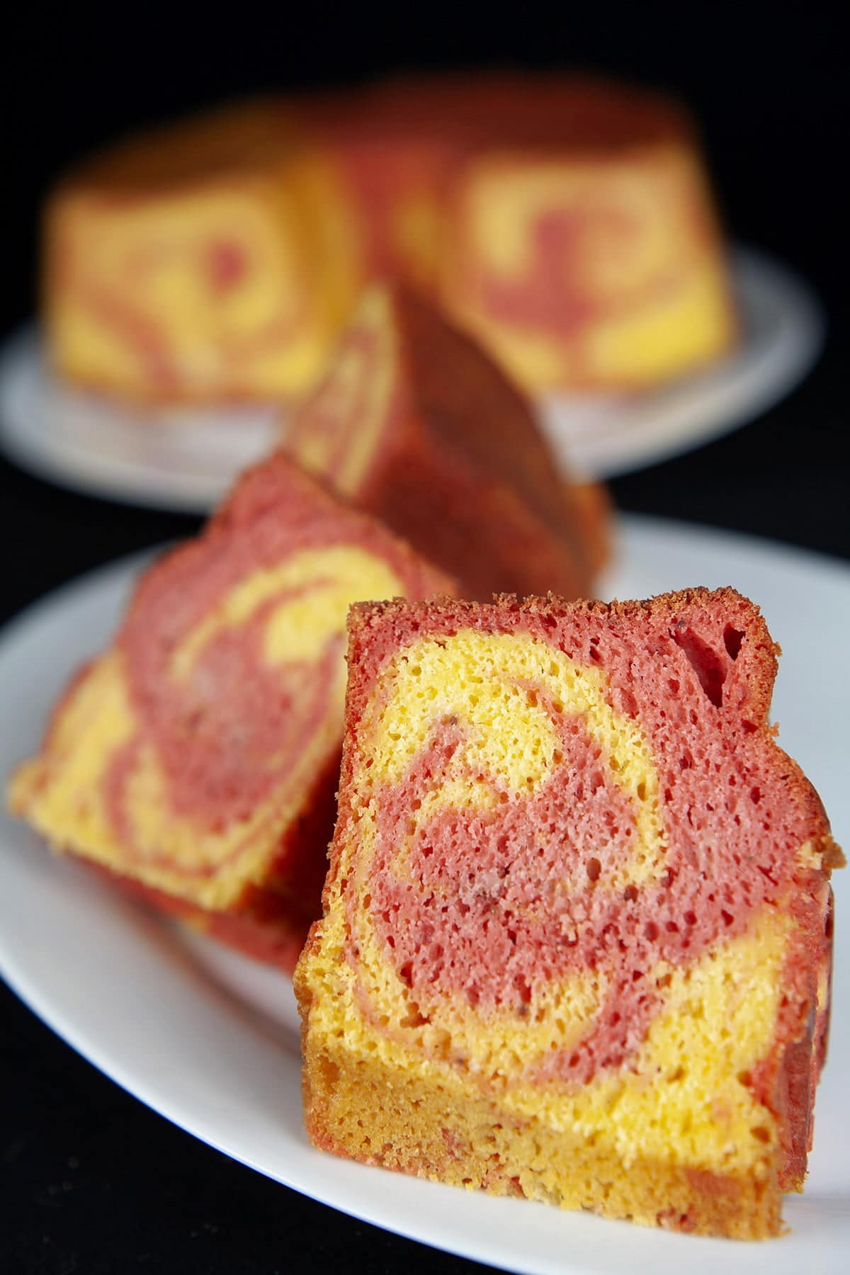 A plate of sliced strawberry mango marble cake slices rests in front of the cake it was cut from. The cake is made of brightly coloured pink and orange batters, marbled together,