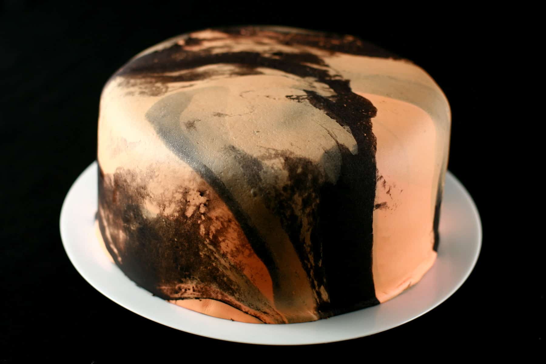 A large round cake on a white plate. The cake is covered with orange and black marbled fondant.