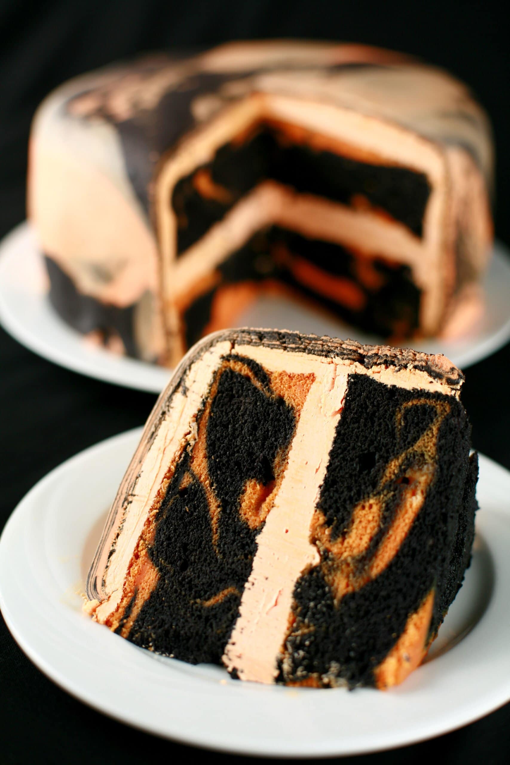 A large round tiger tail cake - black licorice and orange marbled cake, with orange buttercream - is shown with a slcie cut out, and served on a plate in front of it.