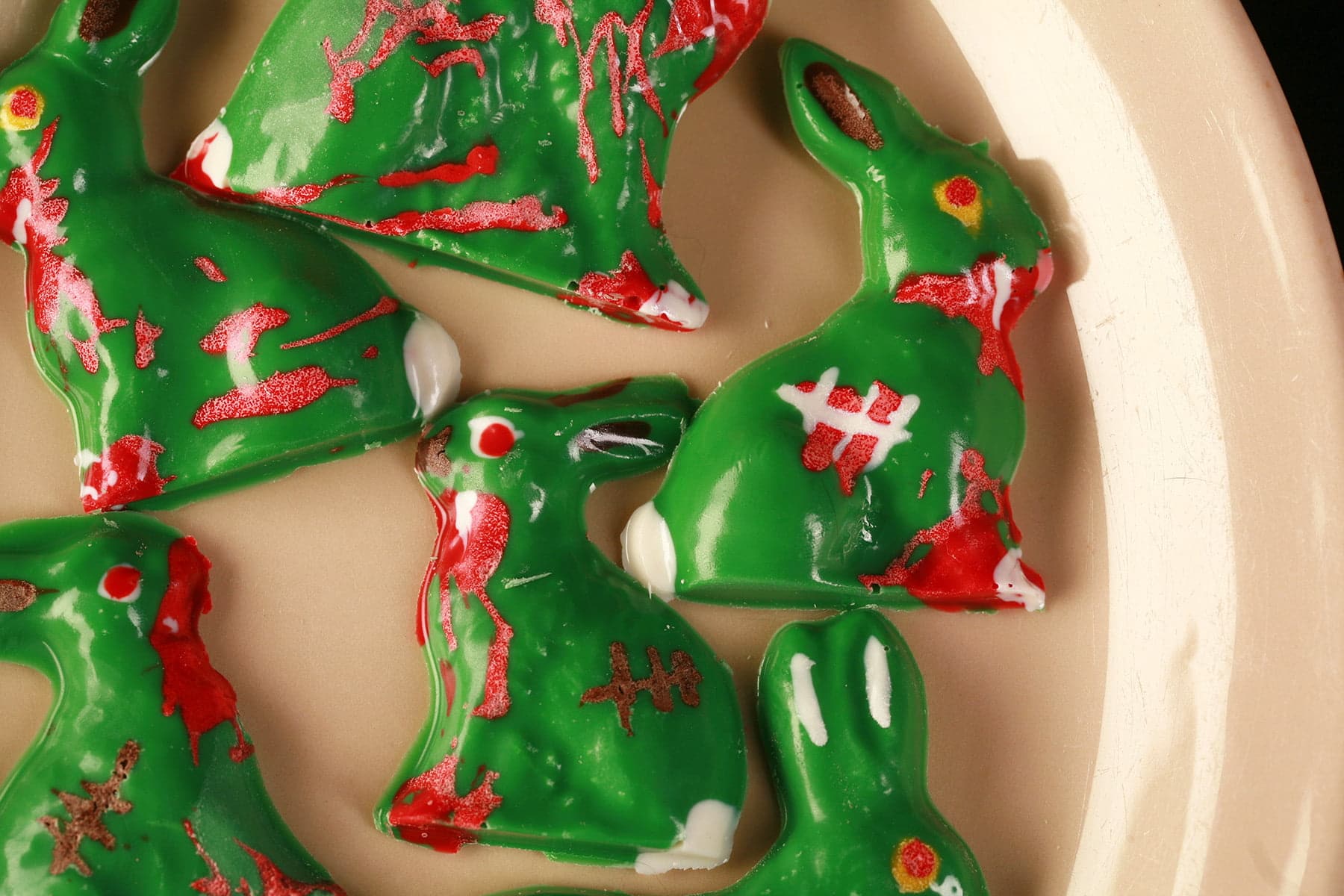6 different Chocolate Zombie Easter Bunnies are arranged on a beige coloured oval plate. They're all green woth red, white, and brown designs to make them look blood spattered, scarred, etc.