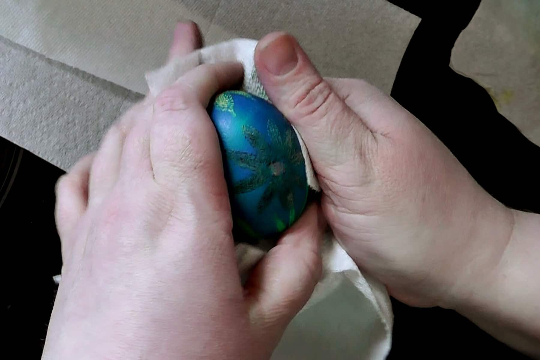 Two hands are shown rubbing melted wax off a pysanky egg. The egg is blue and has daisies on it.