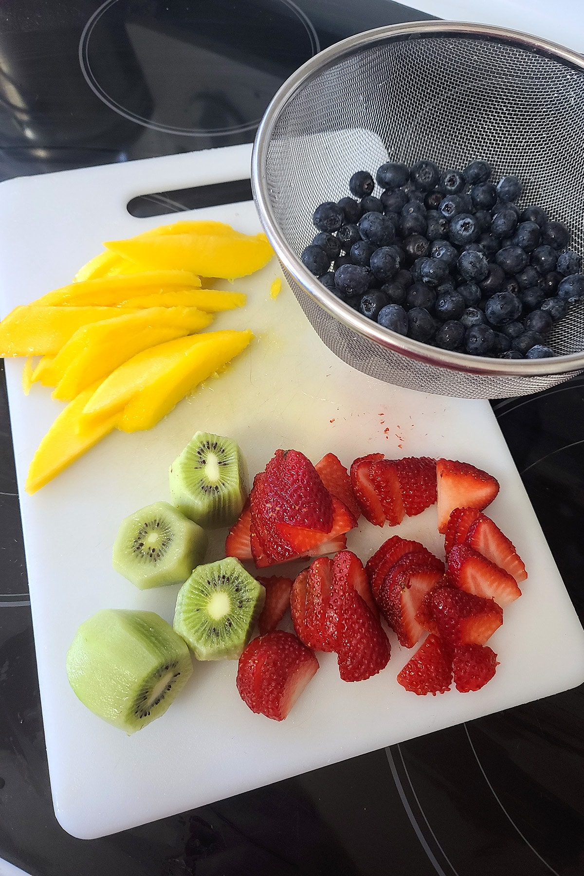 A white cutting board has slices of mango, strawberries, and kiwi fruit, as well as a metal strainer with blueberries in it.