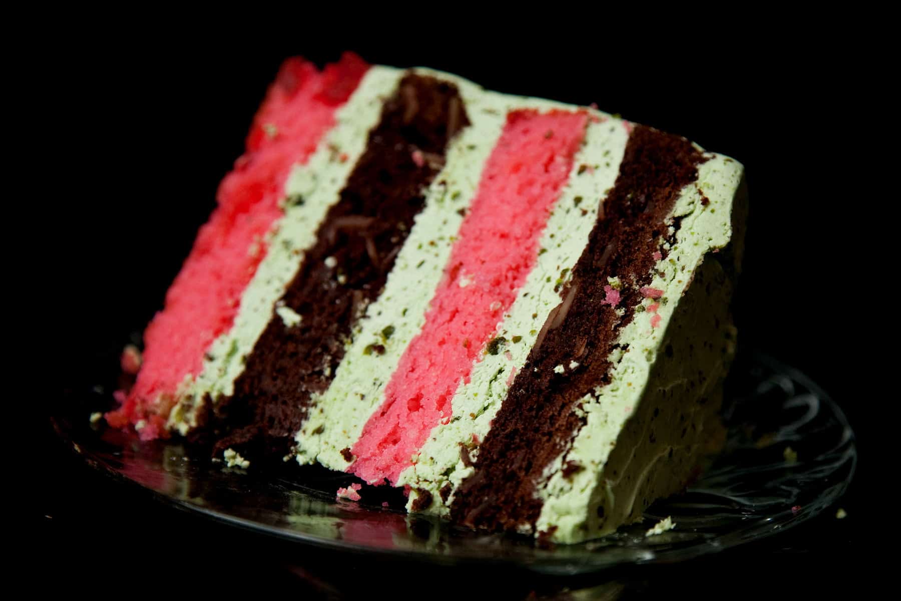 A close up photo of a slice of spumoni torte - alternating layers of chocolate cake and cherry cake, filled and frosted with pistachio buttercream.