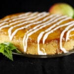 A mango mojito upside down cake: a large round single layer mango upside down cake, on a glass plate. It has a white icing drizzled over it, and has a sprig of fresh mint on the side of the plate.