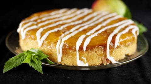 A mango mojito upside down cake: a large round single layer mango upside down cake, on a glass plate. It has a white icing drizzled over it, and has a sprig of fresh mint on the side of the plate.