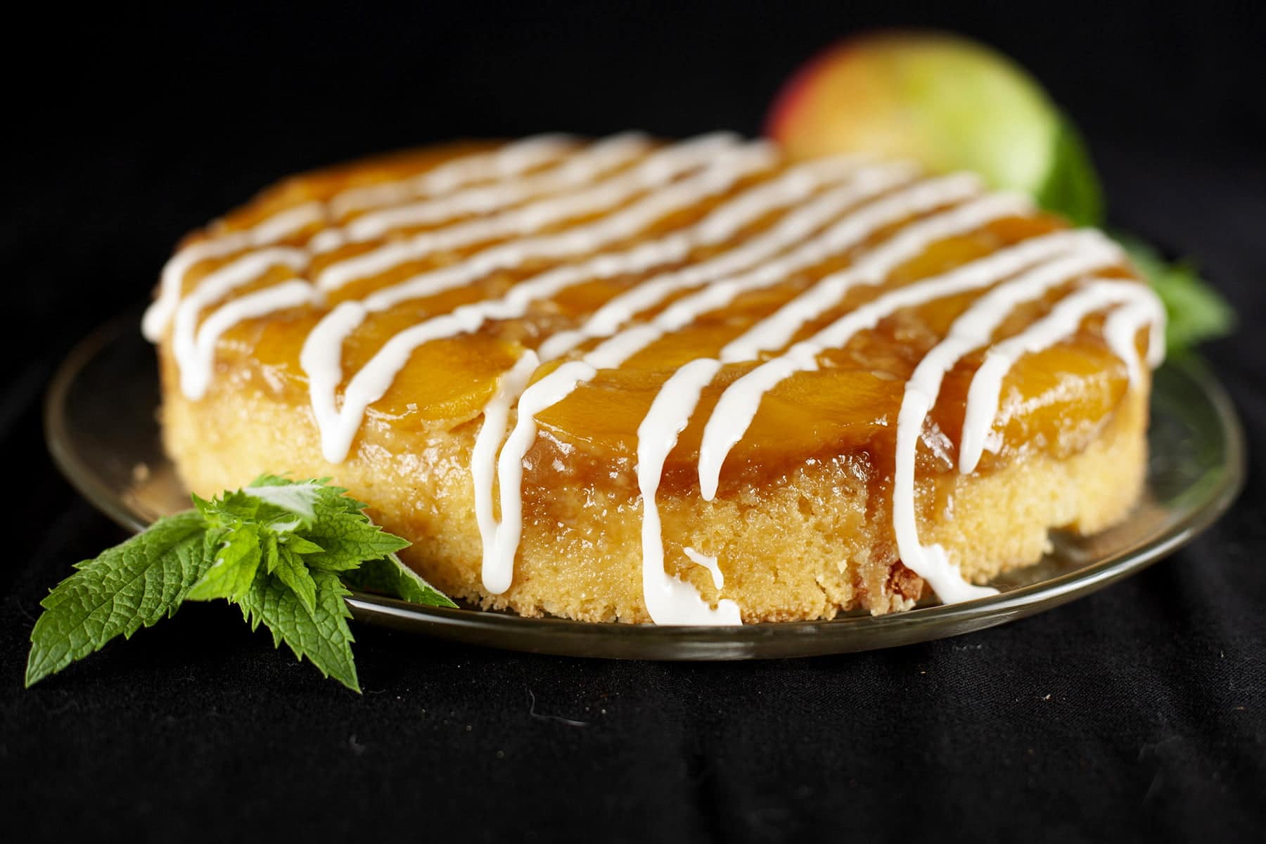A mango mojito upside down cake: a large round single layer mango upside down cake, on a glass plate. It has a white icing drizzled over it, and has a sprig of fresh mint on the side of the plate.