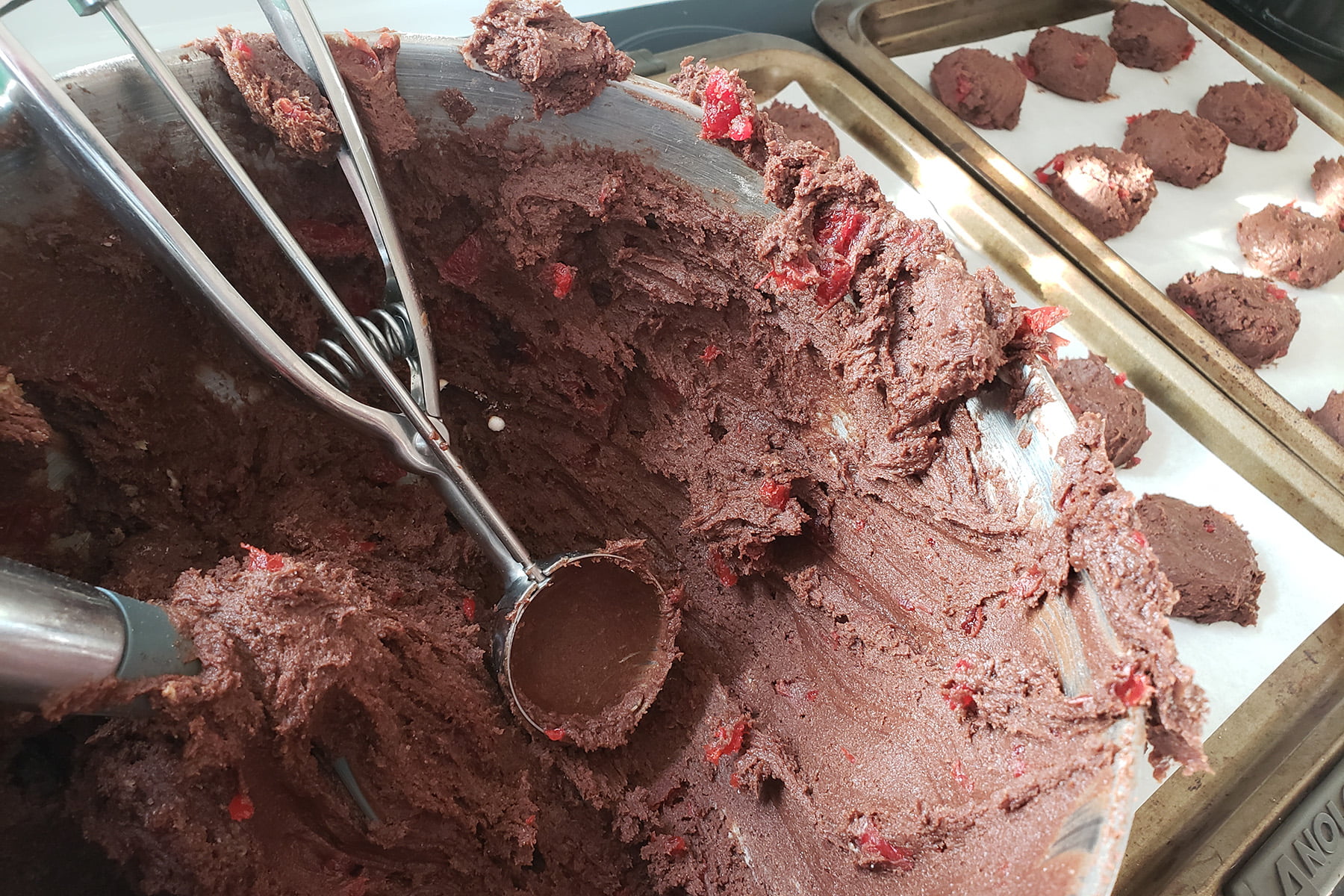 A close up view of a mixing bowl with chocolate cookie dough in it. The dough is generously studded with bits of maraschino cherries. An ice cream scoop rests in the dough.