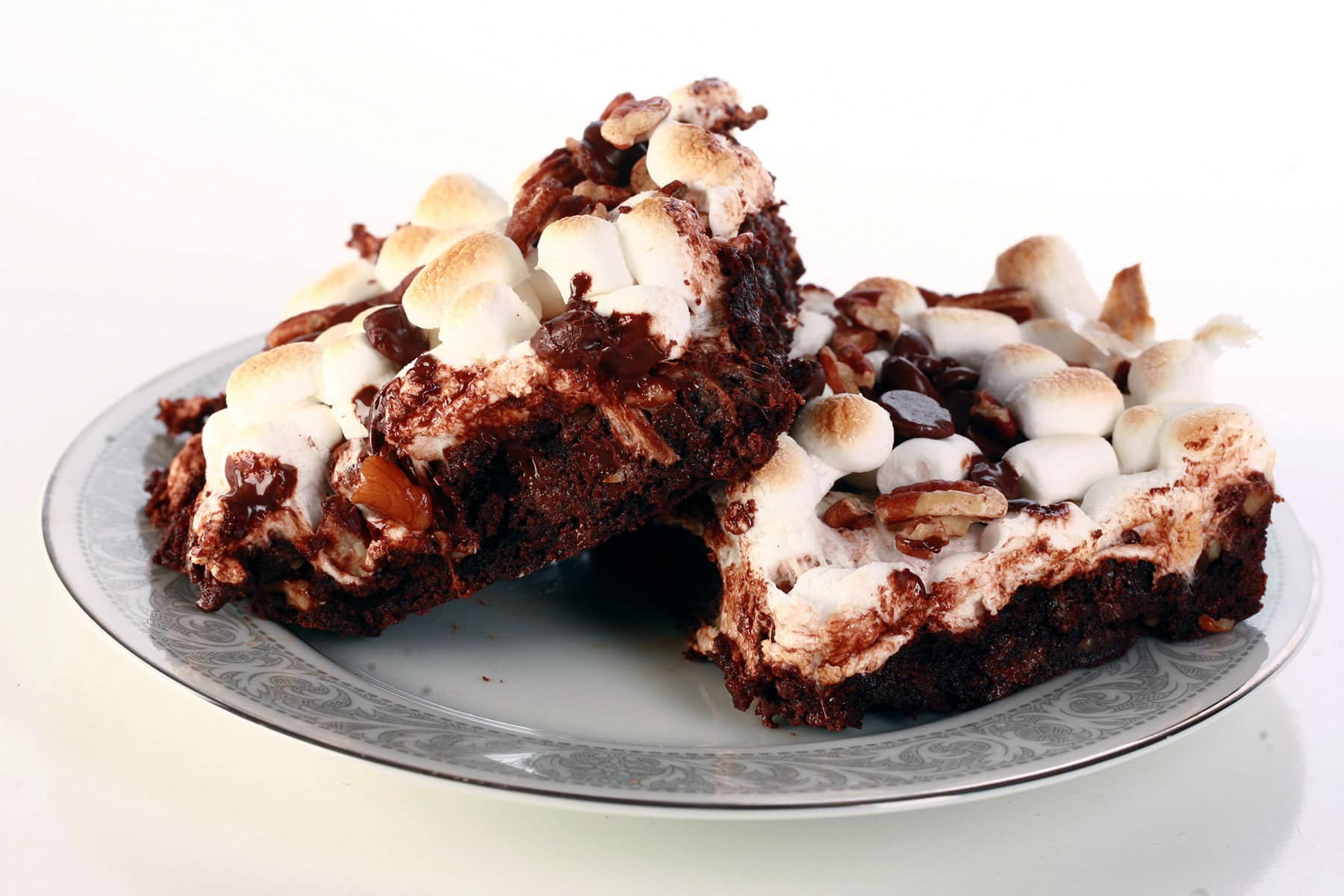 A close up view of two rocky road brownies - double chocolate walnut brownies with toasted mini marshmallows, chocolate chips, and chopped pecans on top.