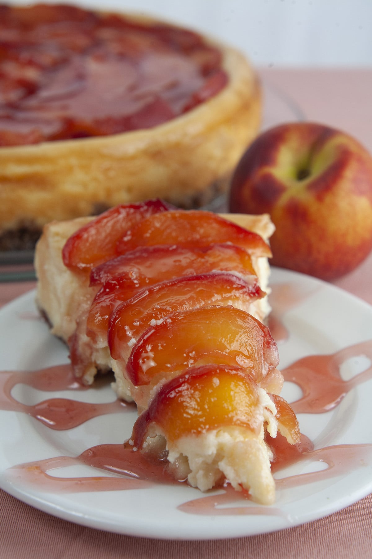 A slice of peach cheesecake with a pink drizzle, next to the whole cheesecake, and a fresh peach.