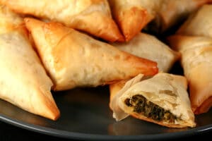 A small blue plate is piled with flaky Spanakopita Triangles. One is cut in half to show the spinach filling.