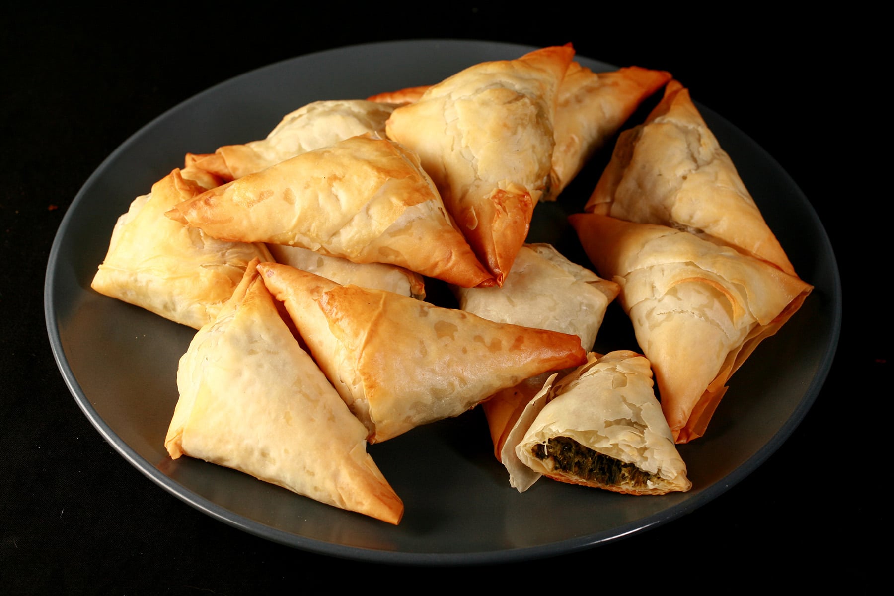 A small blue plate is piled with flaky Spanakopita Triangles. One is cut in half to show the spinach filling.