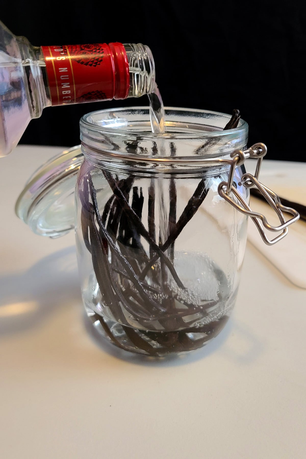 A glass jar with several split vanilla beans in it. Vodka is being poured in.