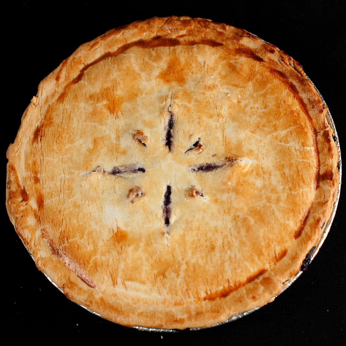 A whole 2-crust pie, against a black background.  Slits cut into the top crust reveal it to be a blueberry pie.